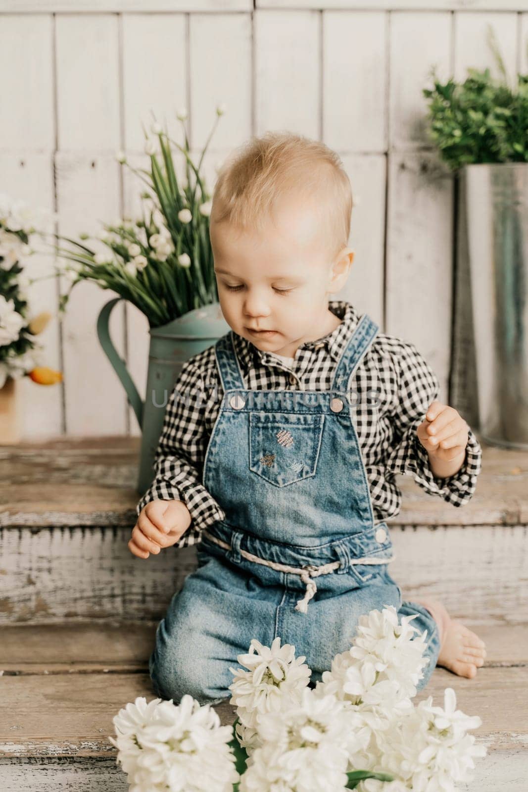 Girl 1 year old. sits holding a white hyacinth with a bulb in her hands, she is dressed in a plaid shirt and denim overalls on a wooden background with green plants in pots