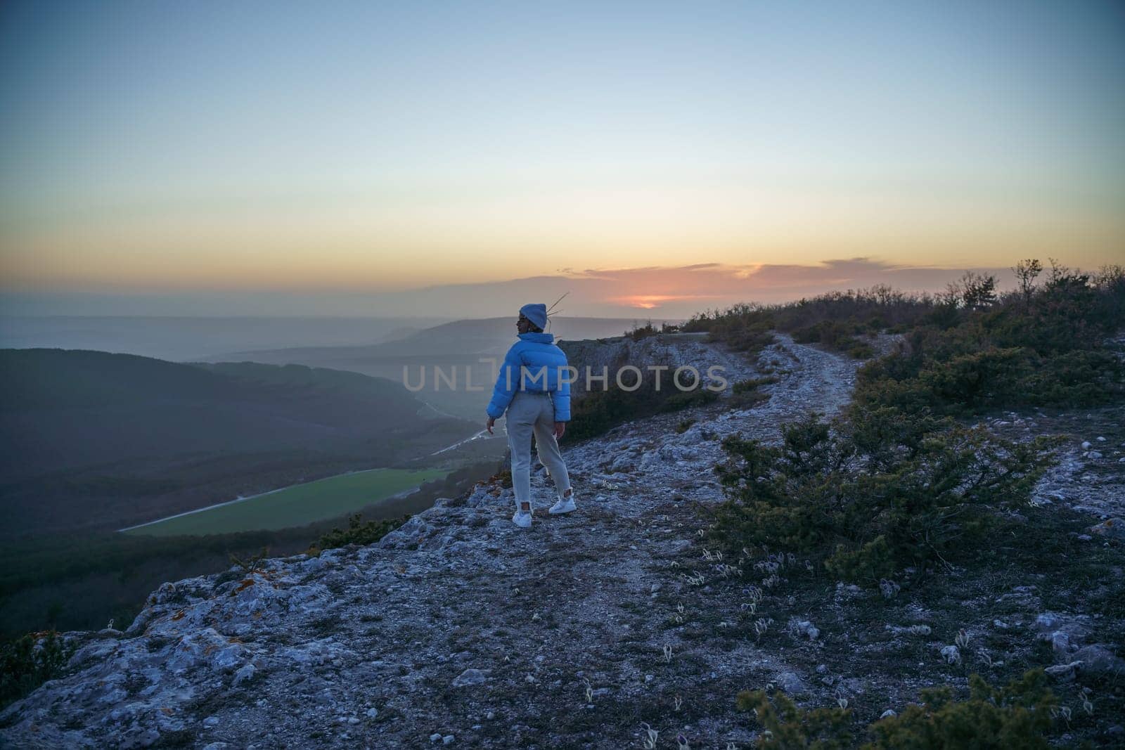Woman tourist on top of sunrise mountain. The girl salutes the sun, wearing a blue jacket, white hat and white jeans. Conceptual design. by Matiunina