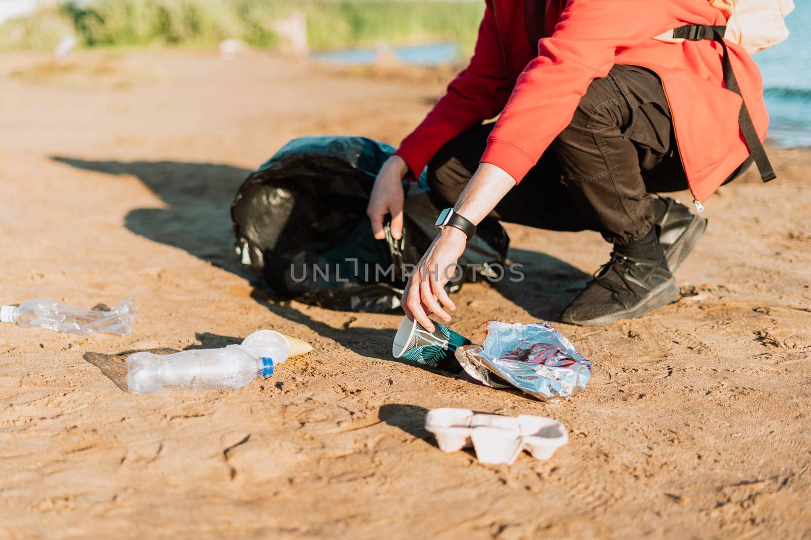 Young male volunteer satisfied with picking up trash, a plastic bottles and coffee cups, clean up beach with a sea. Man collecting garbage. Environmental ecology pollution concept. Earth Day.