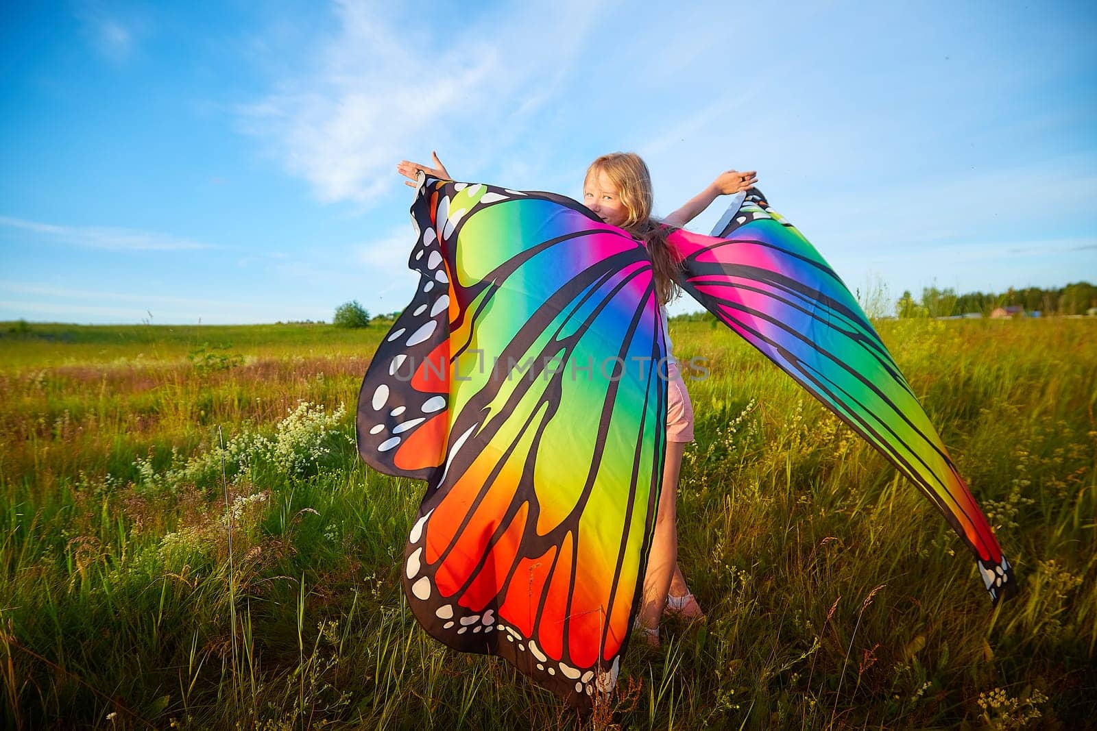 Pretty blonde girl with bright butterfly wings having fun in meadow on natural landscape with grass and flowers on sunny summer day. Portrait of teenage child in spring season outdoors on field