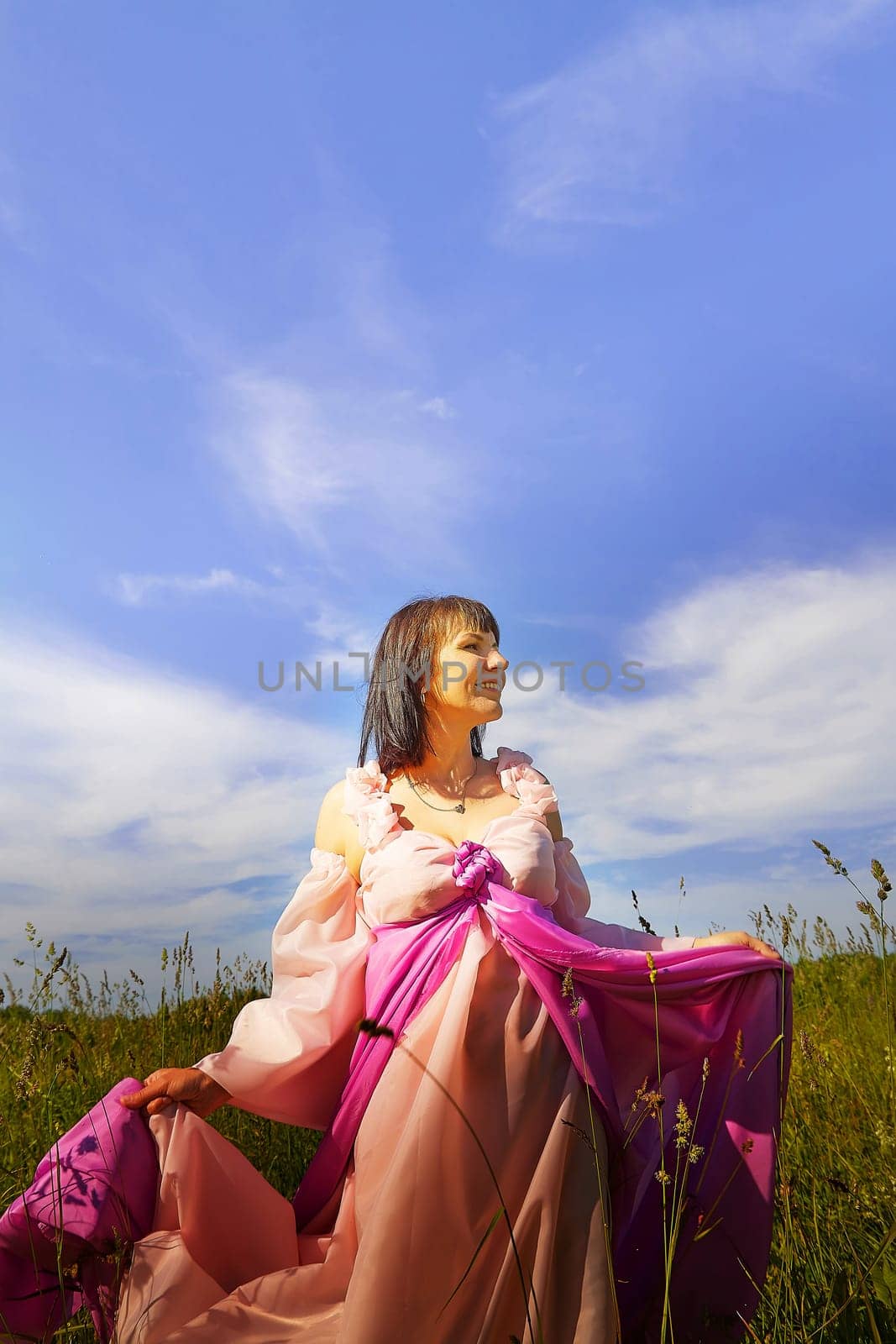 Beautiful girl in lush pink ball gown in green field during blooming of flowers and blue sky on background. Model posing on nature landscape as princess from fary tale