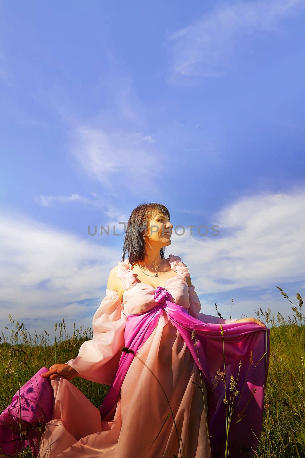 Beautiful girl in a lush pink ball gown in green field during blooming of flowers and blue sky on background. Model posing on nature landscape as princess from fary tale by keleny