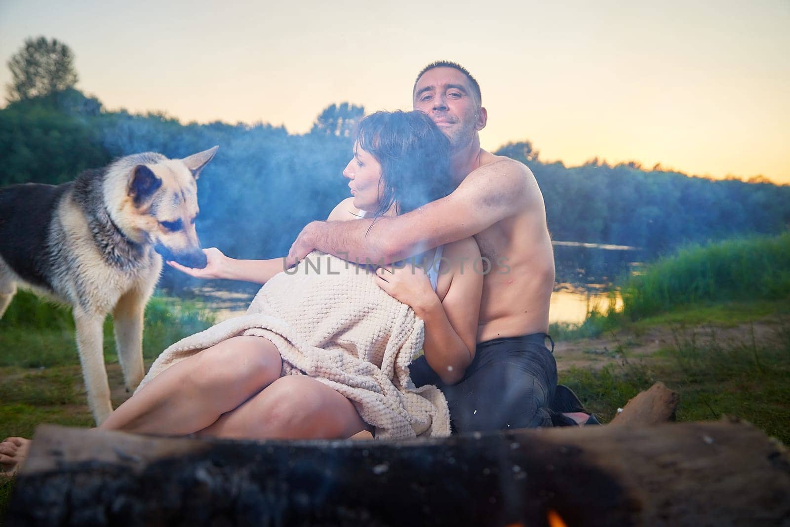 Happy wet couple relaxin, having fun and hugs with big dog near fire in camping on nature at summer sunny evening in sunset. Family or lovers have date and rest outdoor. Concept of love by keleny