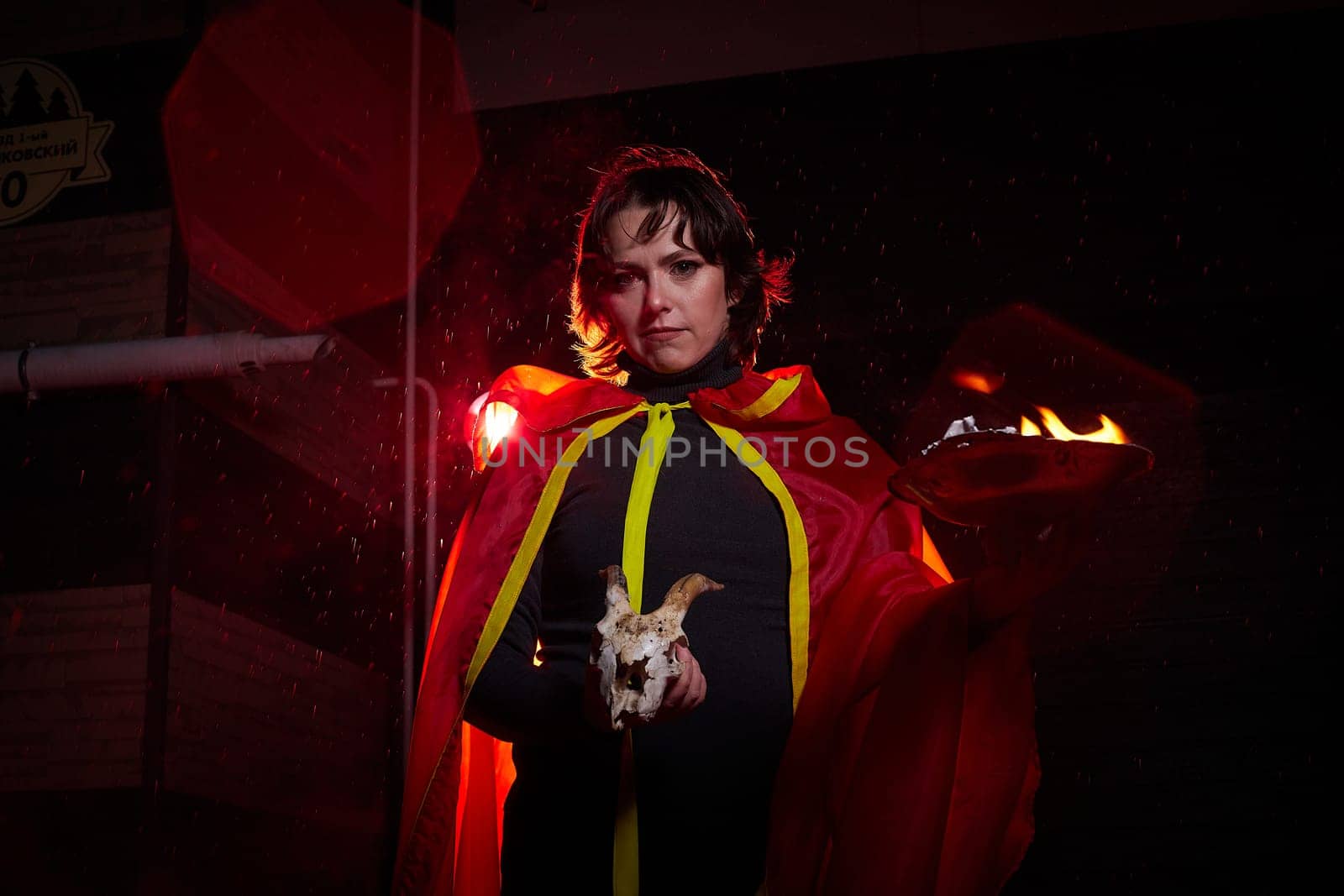 Witch woman in red cloak with skull on black background and light in night. Female warrior in Dark
