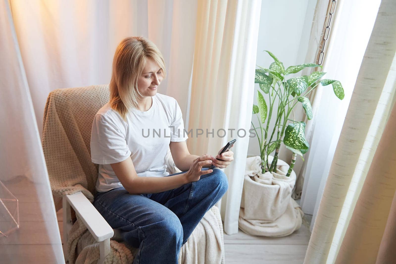 Young blonde woman using cell phone at home. Girl is resting, chating and looking in camera on smartphone near window at home. Nice atmosphere. The concept of cozy home and communication by Internet