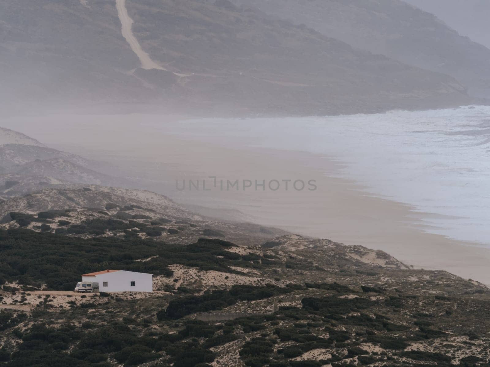 A serene and tranquil scene featuring a solitary house standing on the shores of the Atlantic Ocean in Portugal. The subdued colors and misty atmosphere created by the crashing waves provide a feeling of calmness. Adjacent to the house is a van