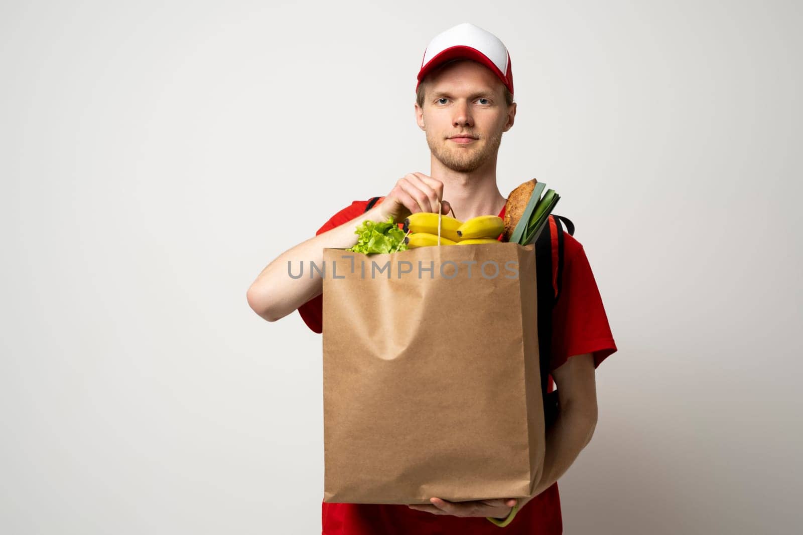 Happy delivery man in red uniform with cap and thermal bag backpack holds brown craft paper bag with food products isolated on gray background studio. Delivery service from shop restaurant to home