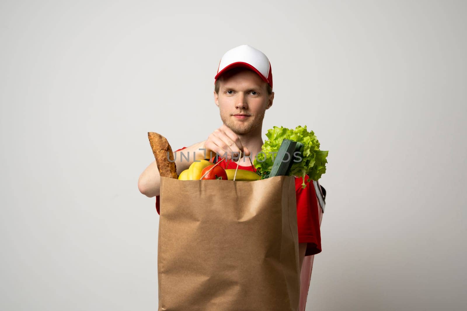 Delivery man employee in red cap and t-shirt uniform hold craft paper packet with food isolated on white background studio