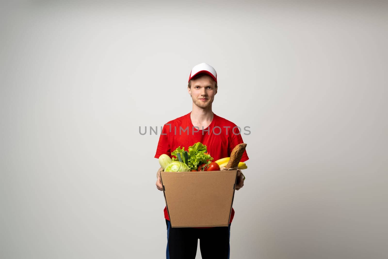 Delivery Concept. Smiling young delivery man in red uniform carrying package box of grocery food and drink from store, market on white studio background