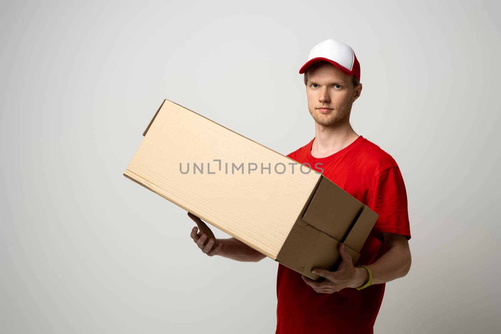 Image of bearded curly delivery man in red uniform with a blank cardboard, percel on white background