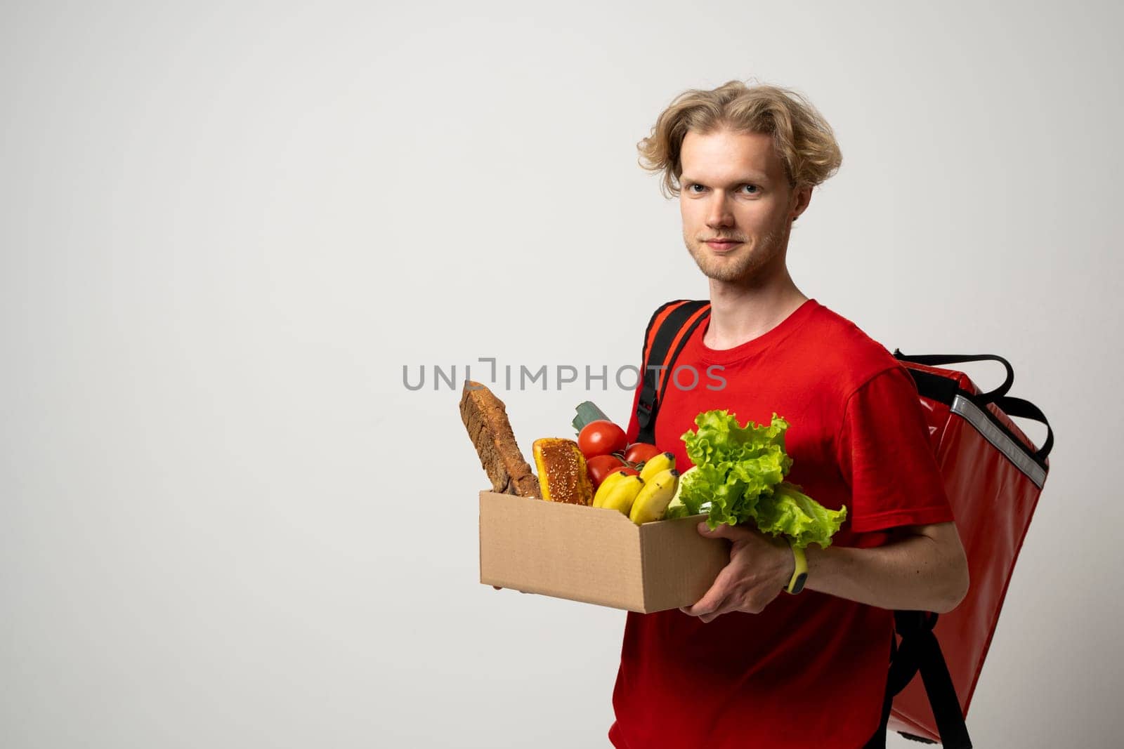 Happy delivery man in red uniform with a box full of groceries over white background. Express delivery, food delivery, online shopping concept. by vovsht