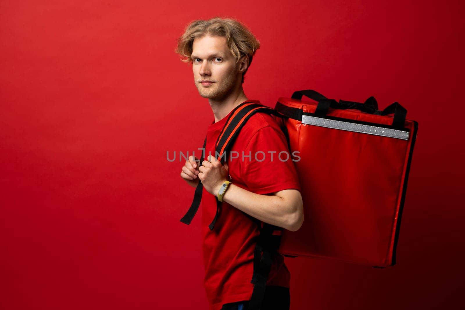 Portrait of young courier with curly hair in red uniform with red thermo bag on red background looks straight in camera. Food delivery service