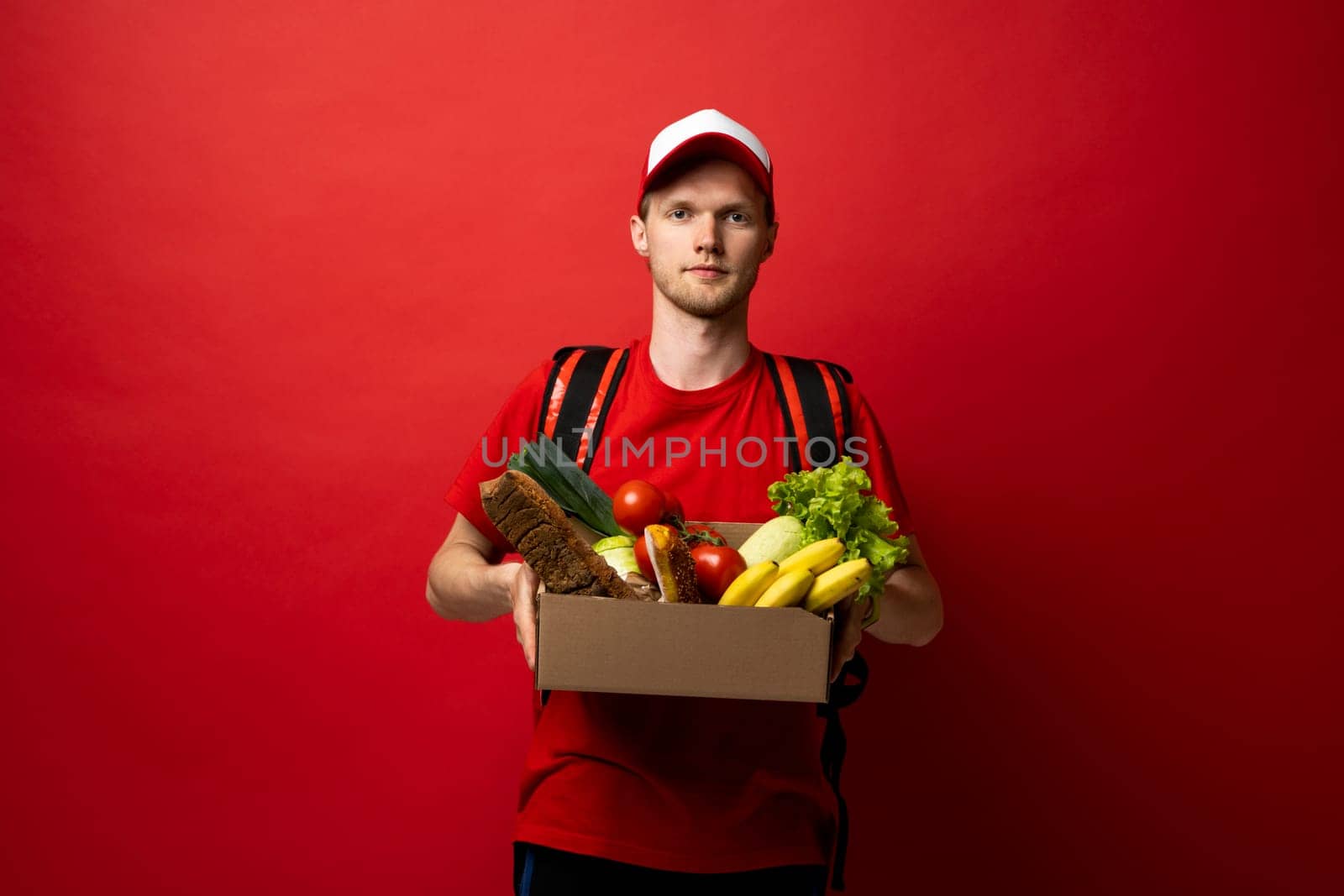 Courier in red uniform holds paper box of food, groceries. Delivery service, takeaway restaurants food delivery to home door