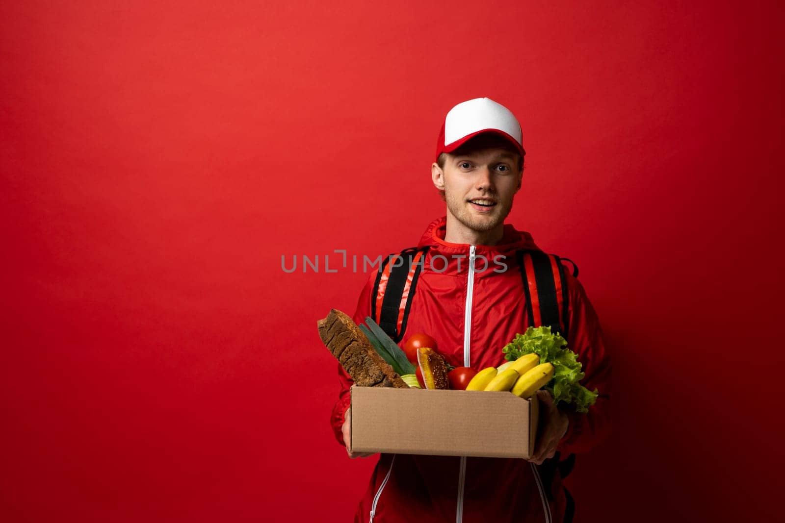 Delivery man in red uniform holds cardboard paper box with food isolated on white background, studio portrait. Service concept