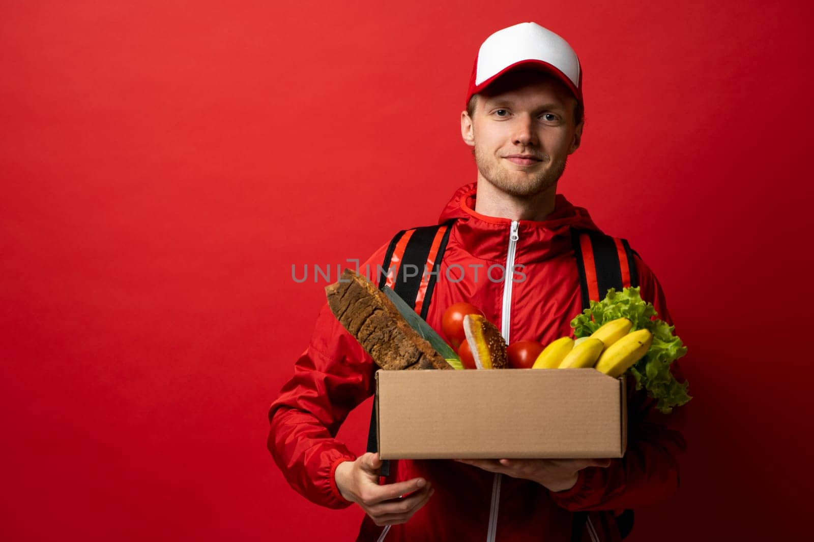 Delivery man in a red uniform holding paper box full of groceries. Online shopping and express delivery. Quality service of a restaurant