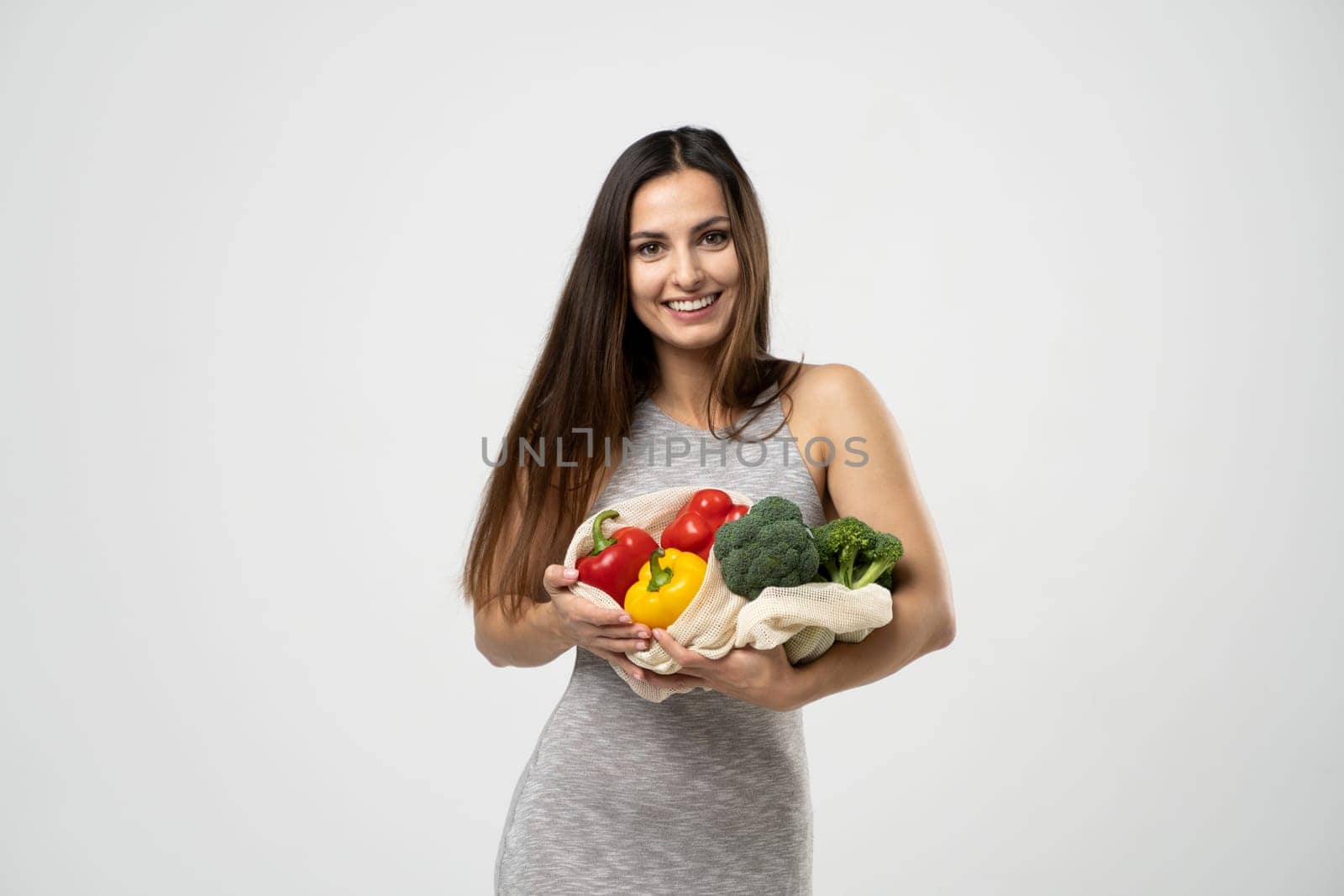 Smiling brunette girl is holding mesh shopping bag with vegetables, greens without plastic bags. Zero waste, plastic free Eco friendly concept. Sustainable lifestyle