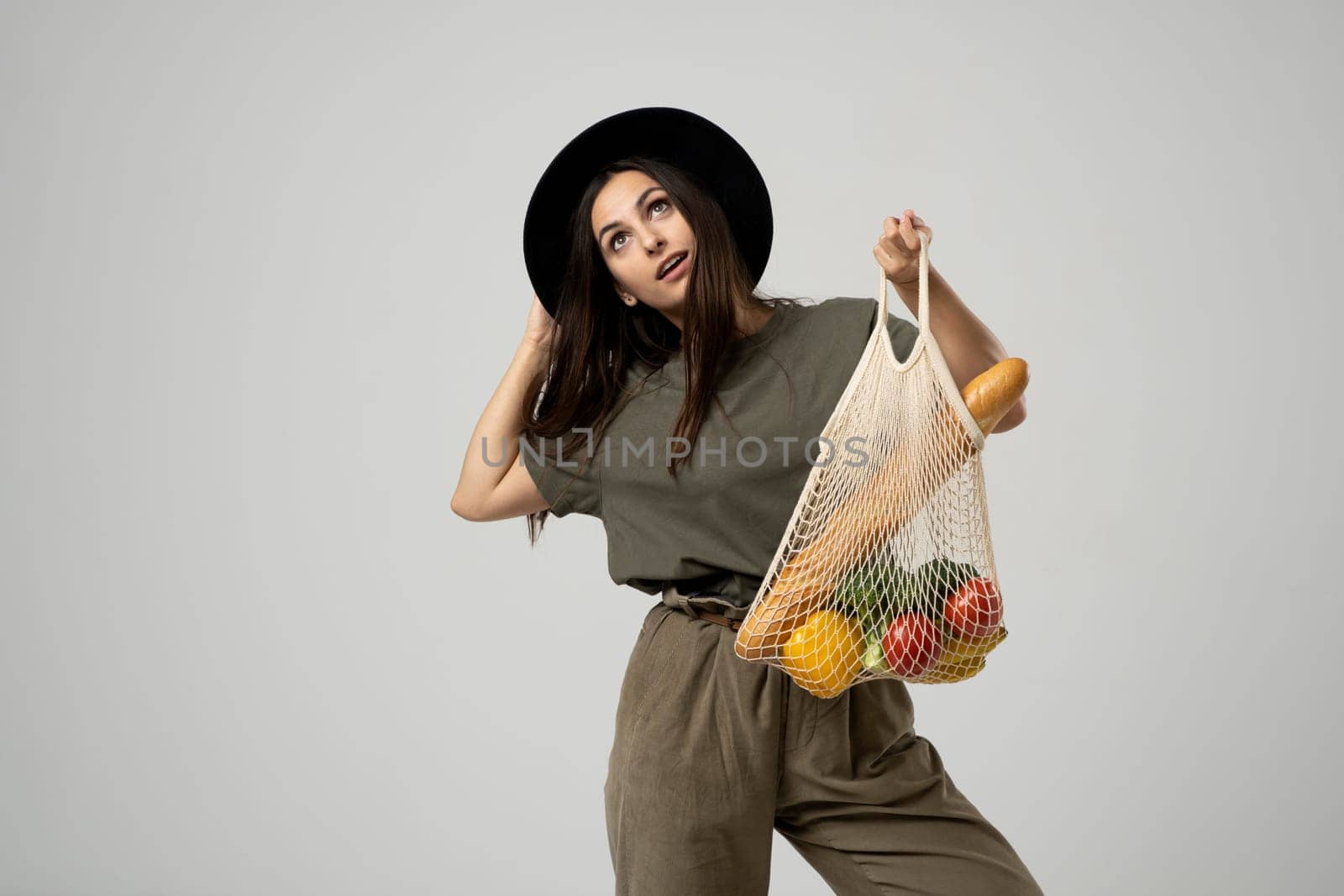 A young fashion brunete girl in a beige t-shirt and black hat holds a mesh reusable eco bag with food, vegetabes, fruits. Zero waste, plastic free. Eco friendly concept. Sustainable lifestyle. by vovsht