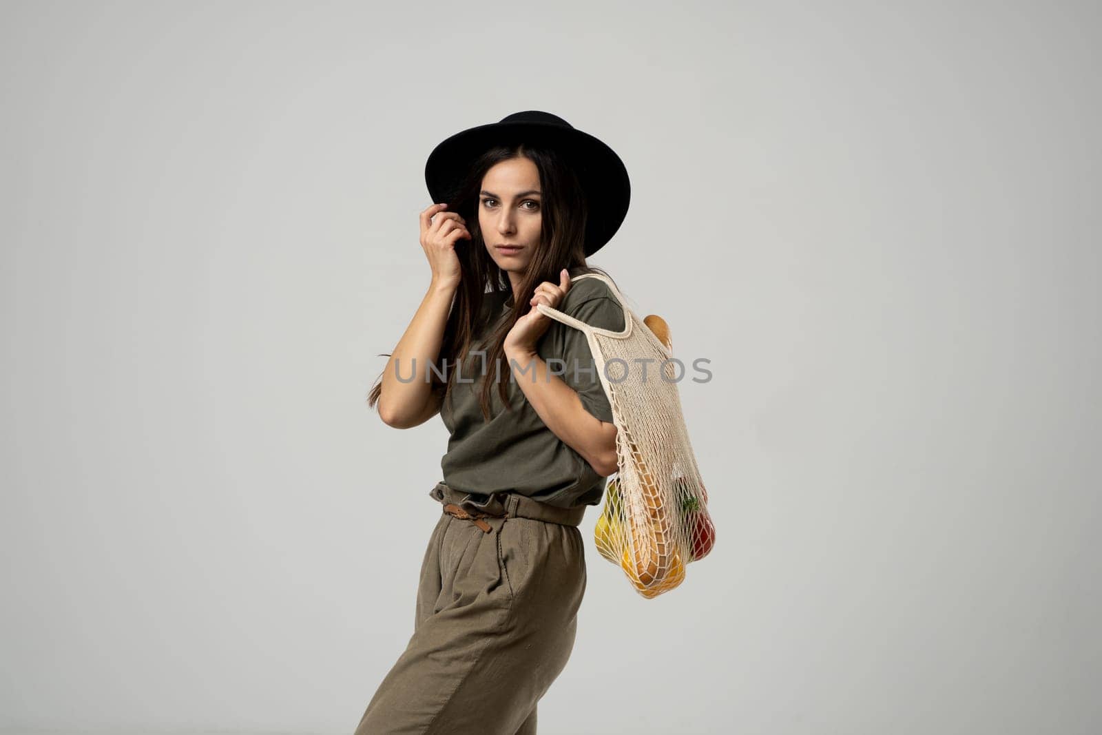 A modern stylish woman with a mesh shopping bag with vegetables looking in camera against the background of the studio. Refusal of plastic bags