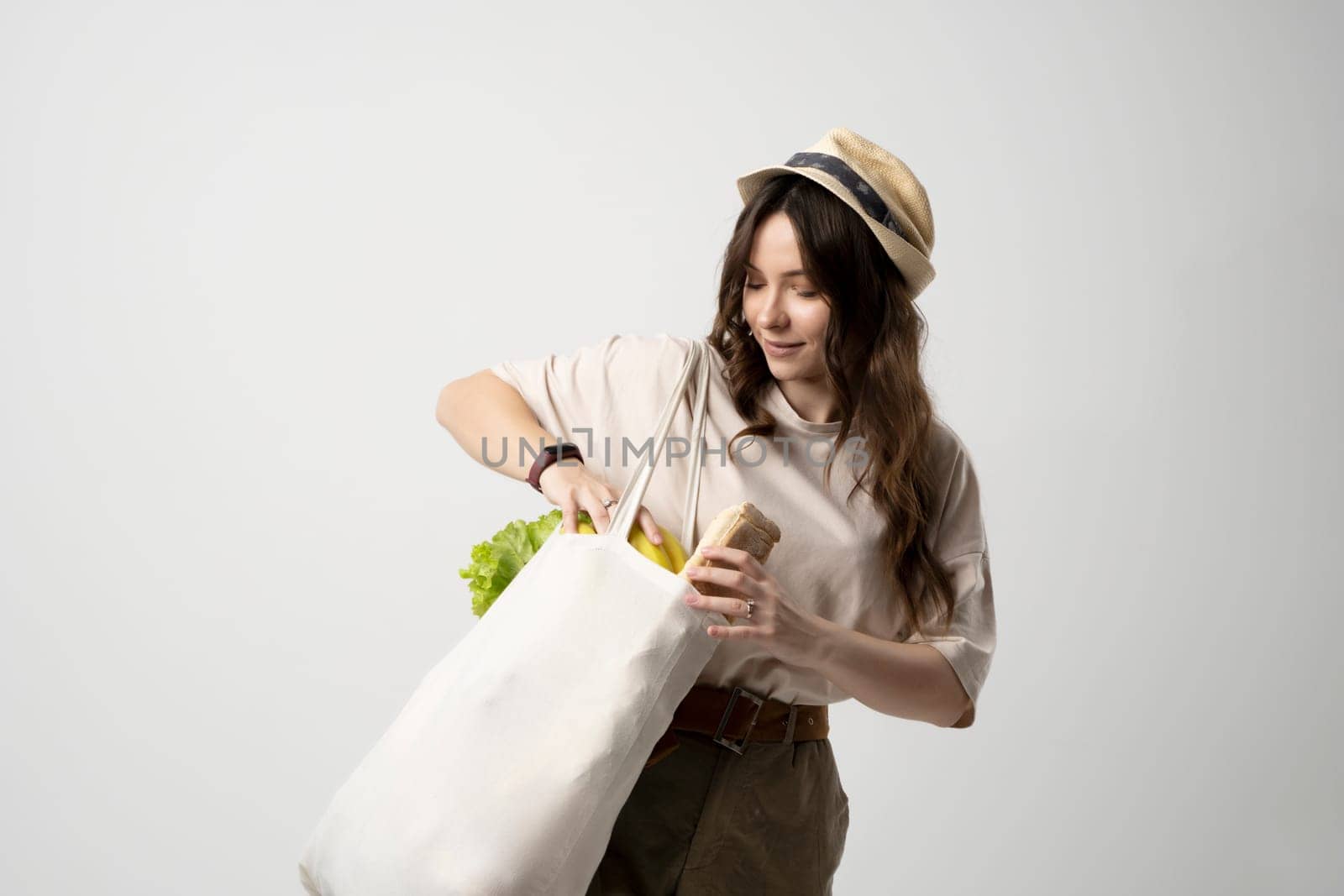 Smiling young woman in beige t-shirt and a hst with a mesh eco bag full of vegetables and on a white studio background. Sustainable lifestyle. Eco friendly concept. Zero waste. by vovsht