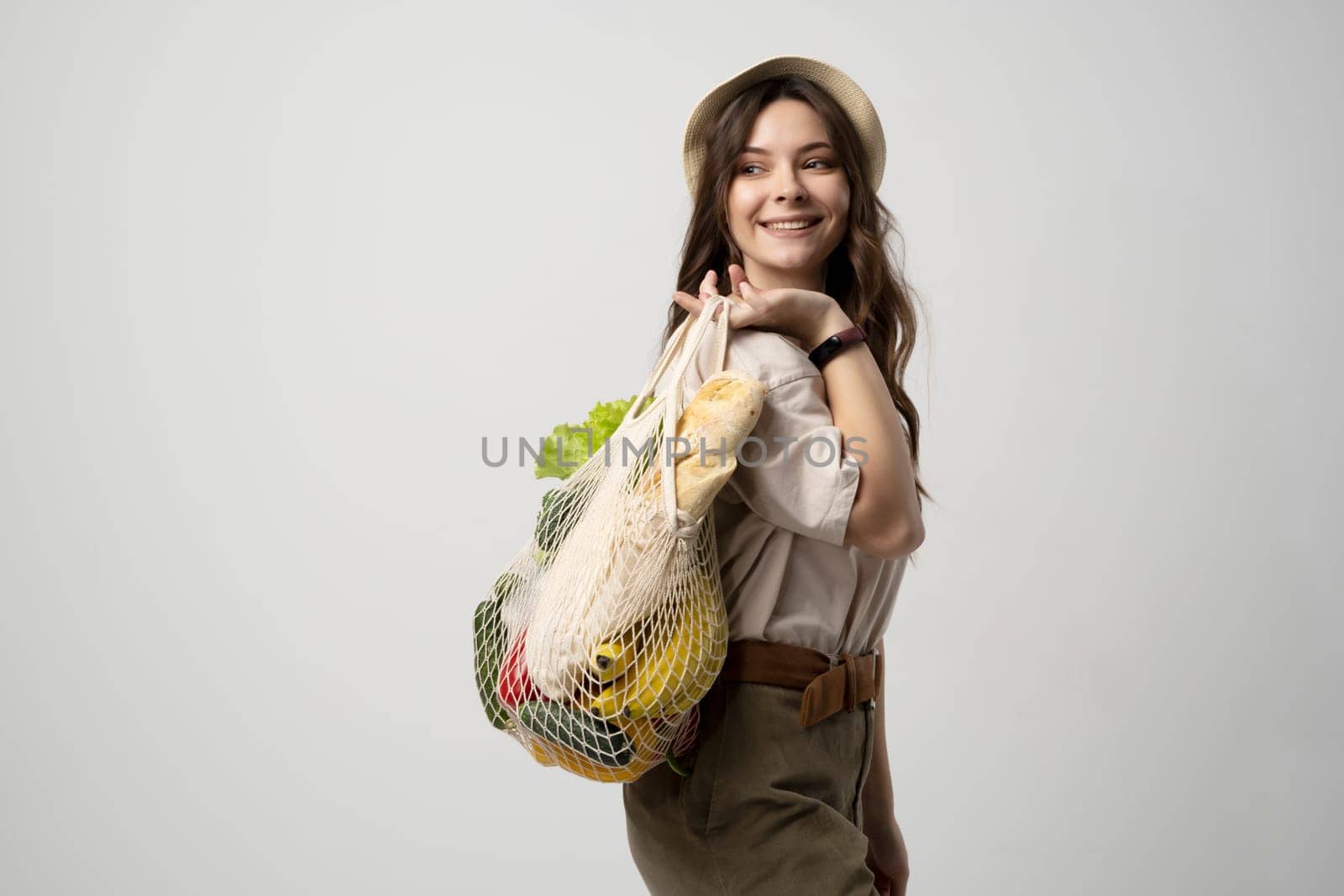 Eco friendly positive young woman in beige oversize t-shirt holding reusable mesh cotton eco bags for shopping with groceries on white background