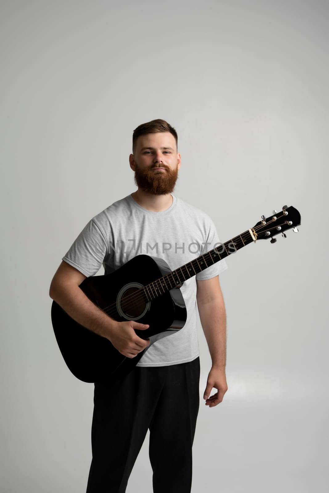 Male musician playing on acoustic guitar. Portraite of bearded handsome musician holding a guitar in a white room. Guitarist, artist, composer, rocker, star