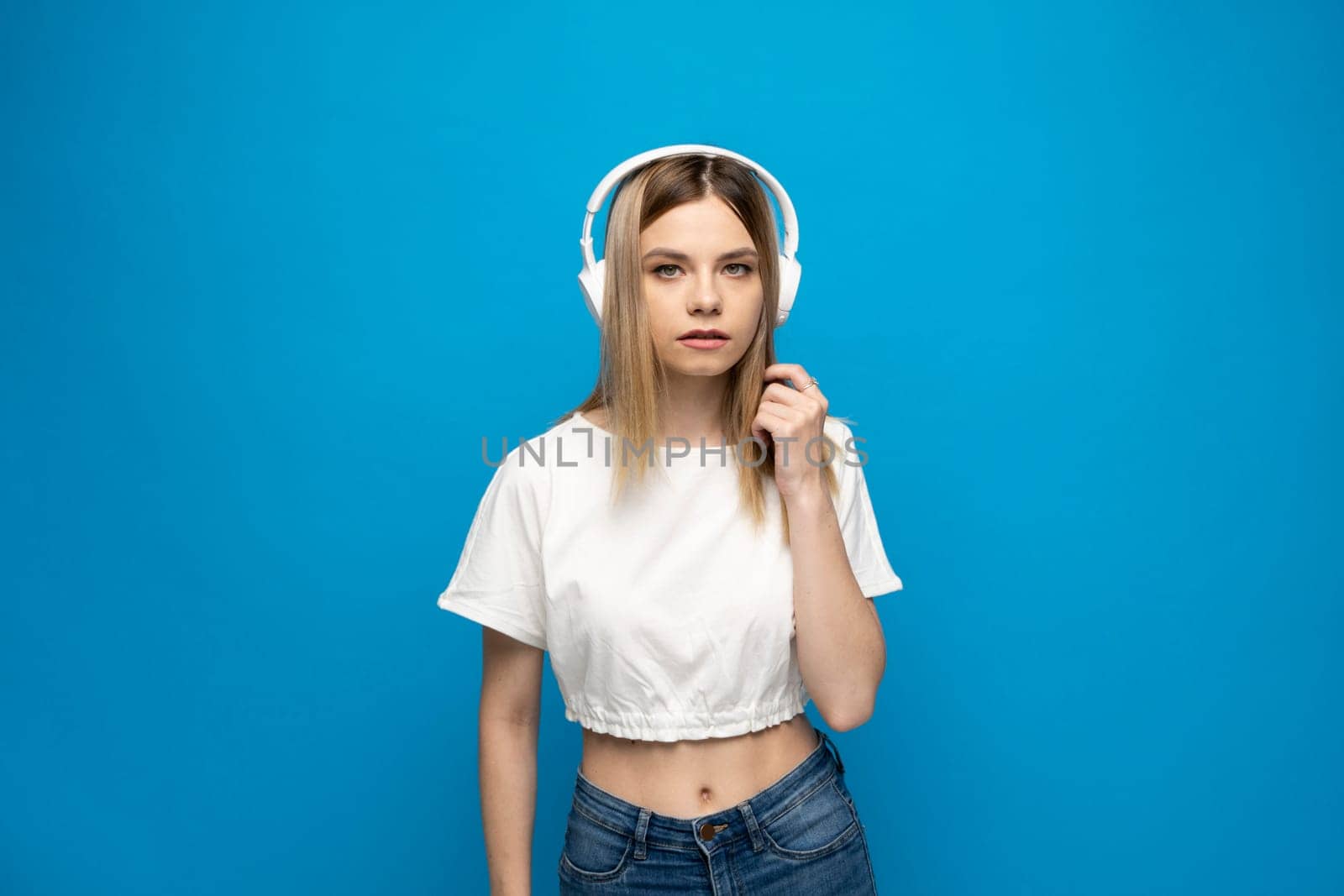 Cheerful young blonde woman girl in white clothes listening music with headphones, mobile phone isolated on blue background studio. People lifestyle concept