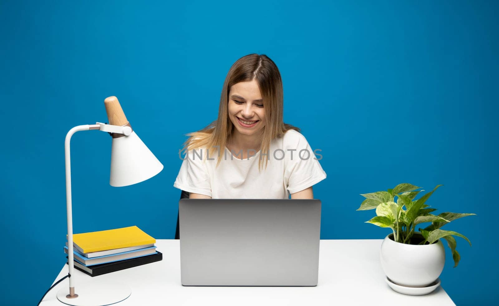Portrait of a pretty young woman studying while sitting at the table with grey laptop computer, notebook. Smiling business woman working with a laptop isolated on a blue background. by vovsht