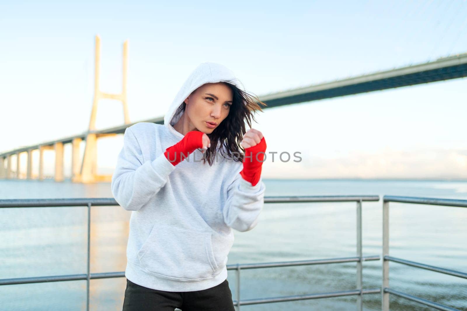 Woman shadow boxing with her hands wrapped in red boxing tapes with ocean bridge and blue sky background. Female athlete fighter training outside dressed hoodie. Copy space