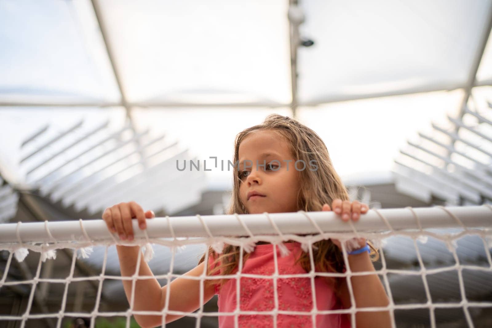 Little girl lean on net looking away with sad face emotion. Lost caucasian child. Female kid unhappy when playground is closed on quarantine