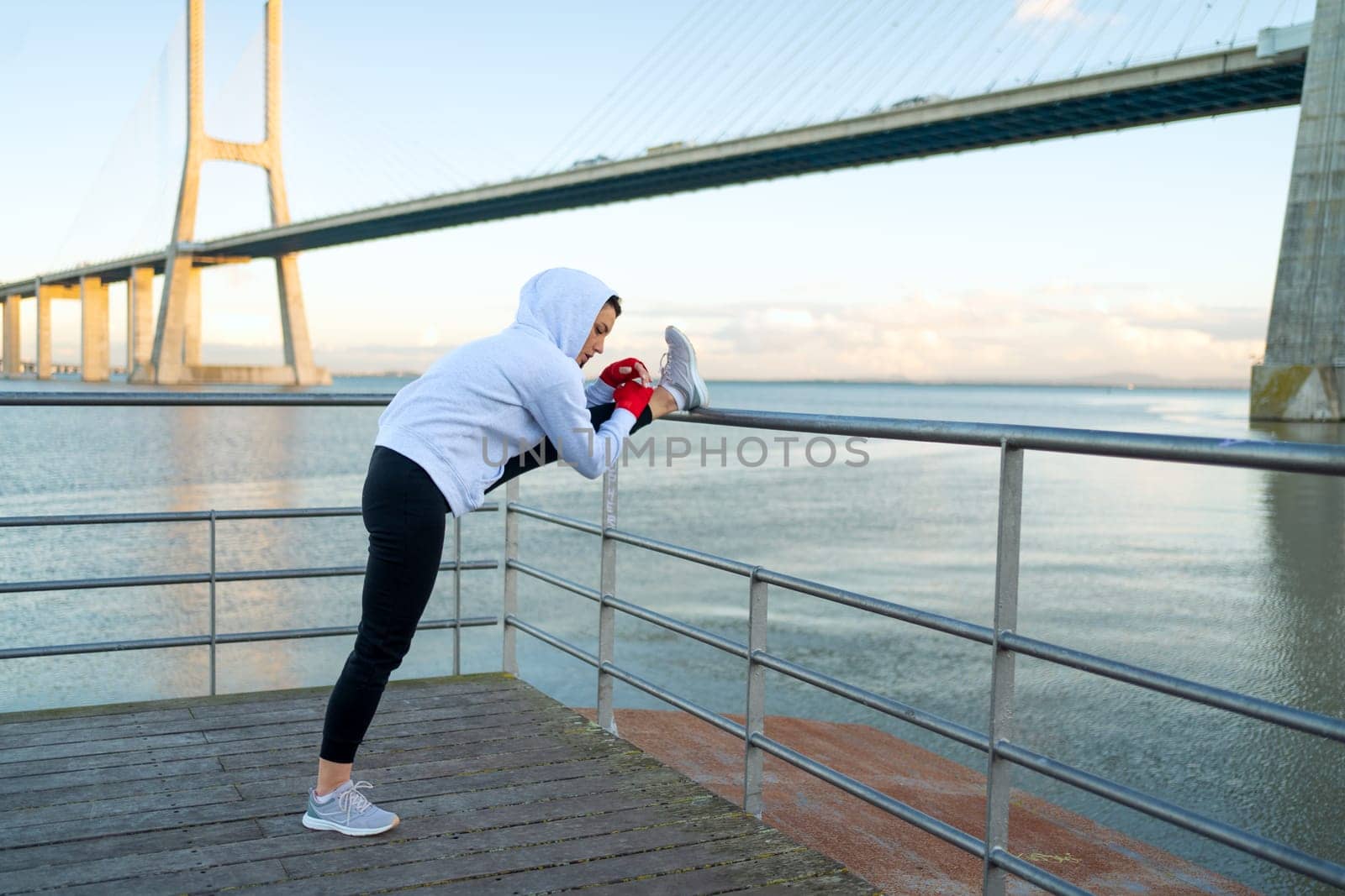 Athlete age woman stretching legs before kickboxing exercises. Caucasian athletic female does lunges exercises stretch leg with bridge and blue sky background. Back view