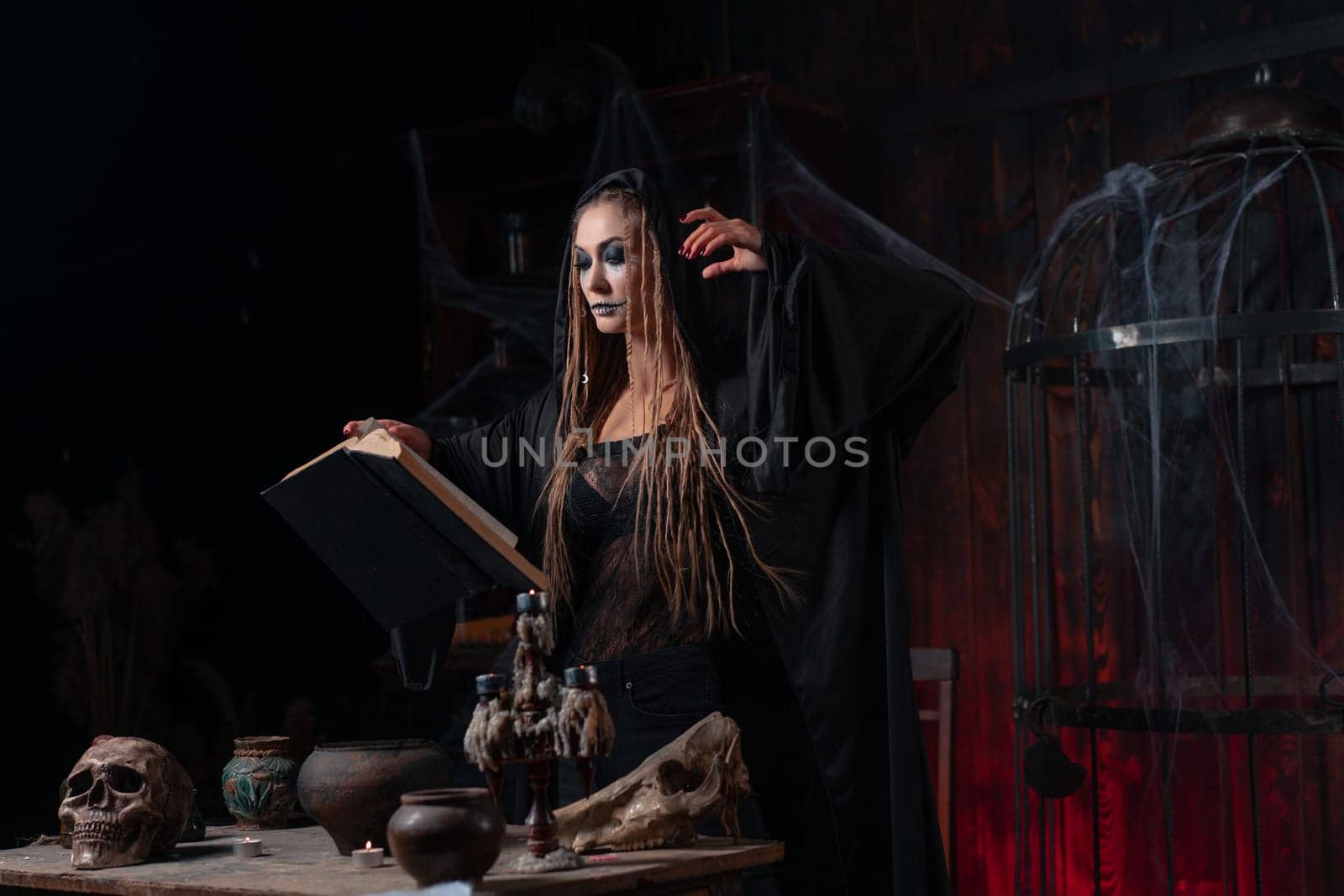 Halloween concept. Witch dressed black hood with dreadlocks standing dark dungeon room use magic book for conjuring magic spell by andreonegin