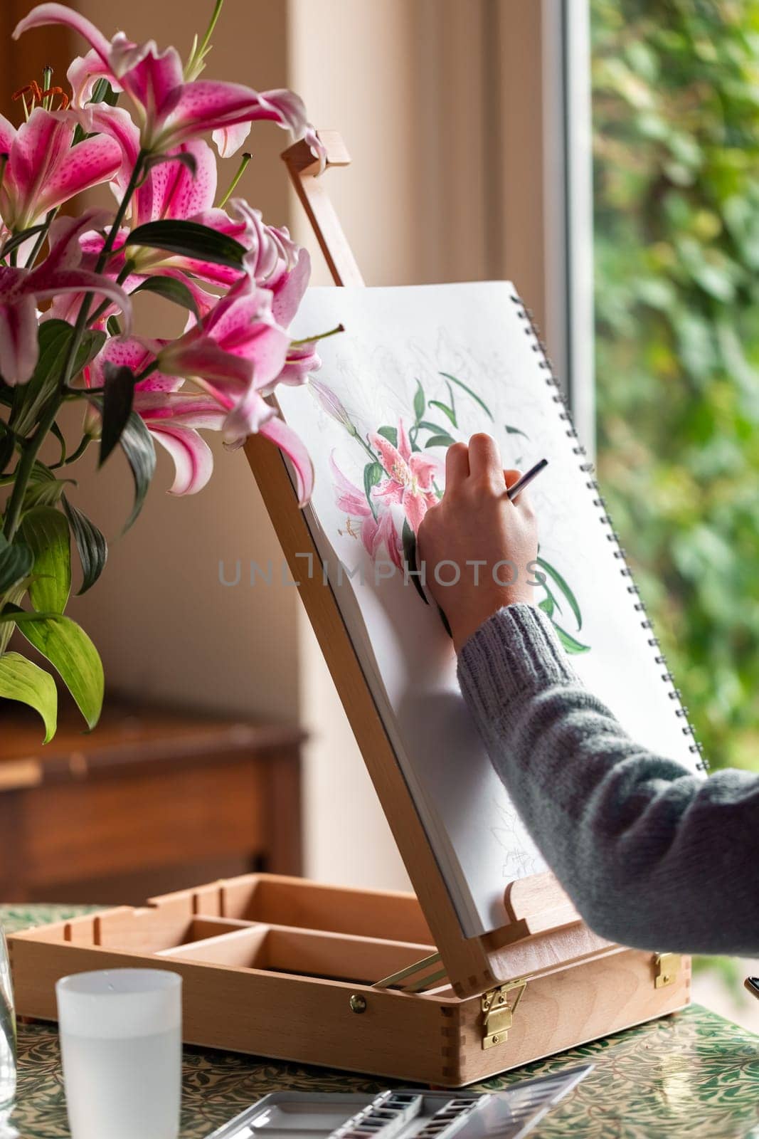 Young left-handed girl painting still life with flowers, purple lilies, with watercolor paints on the easel at home by Len44ik