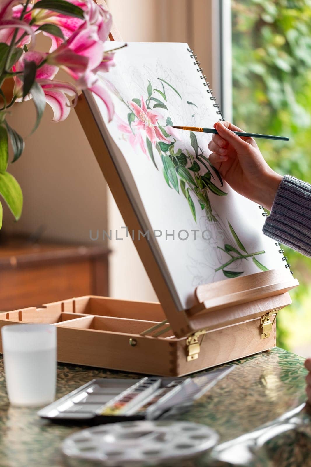 Young girl painting still life with flowers, purple lilies, with watercolor paints on the easel at home