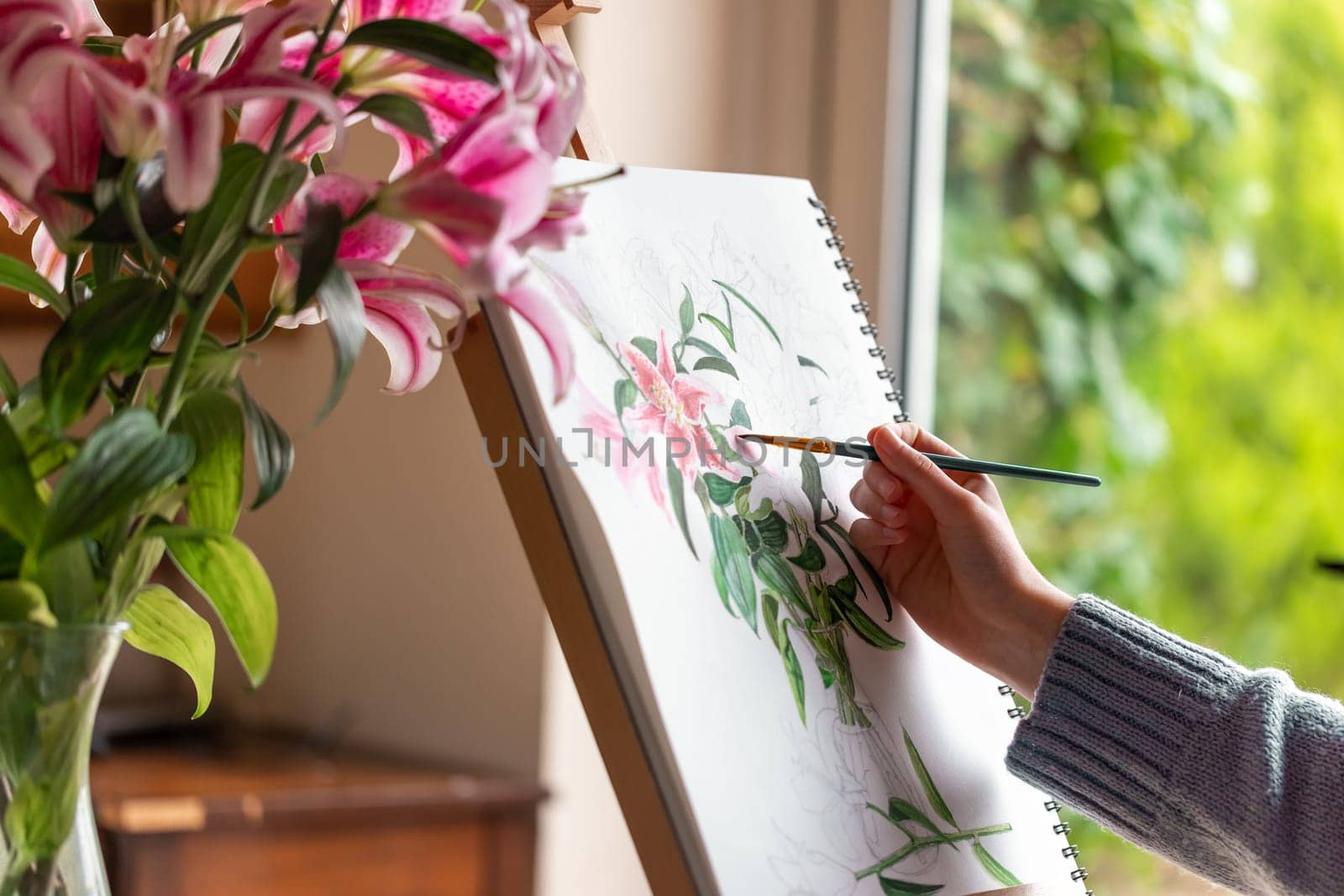 Young girl painting still life with flowers, purple lilies, with watercolor paints on the easel at home