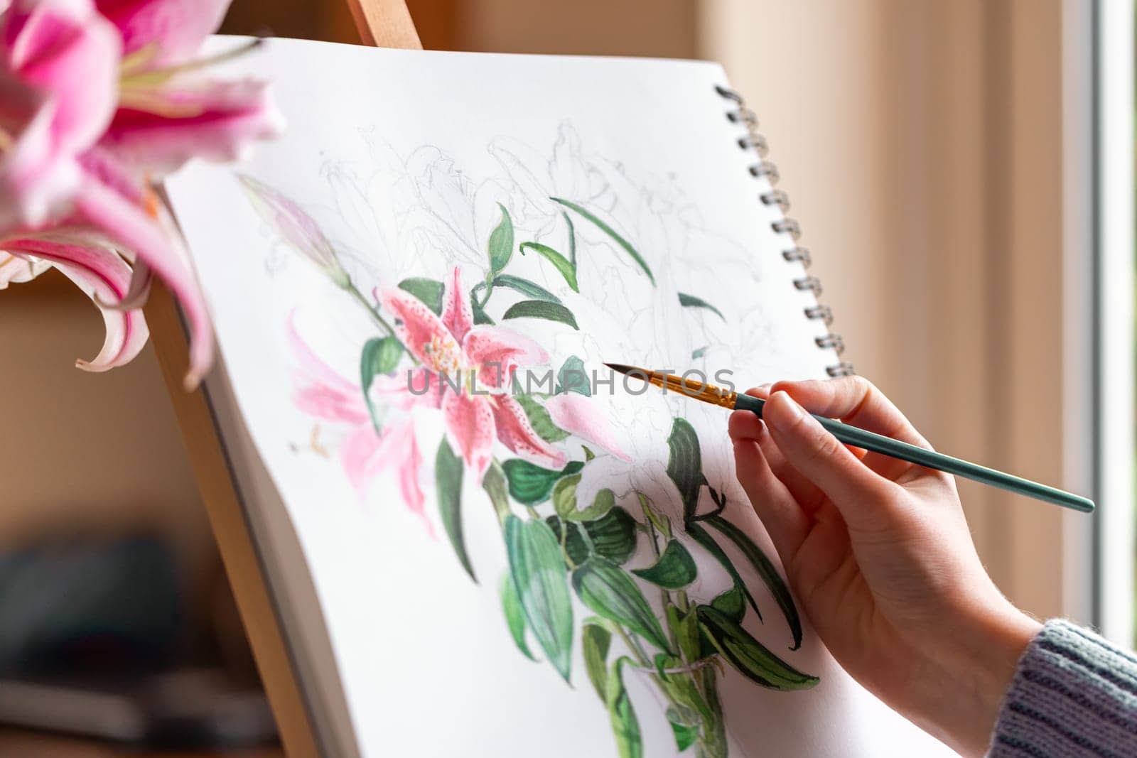 Young girl painting still life with flowers, purple lilies, with watercolor paints on the easel at home