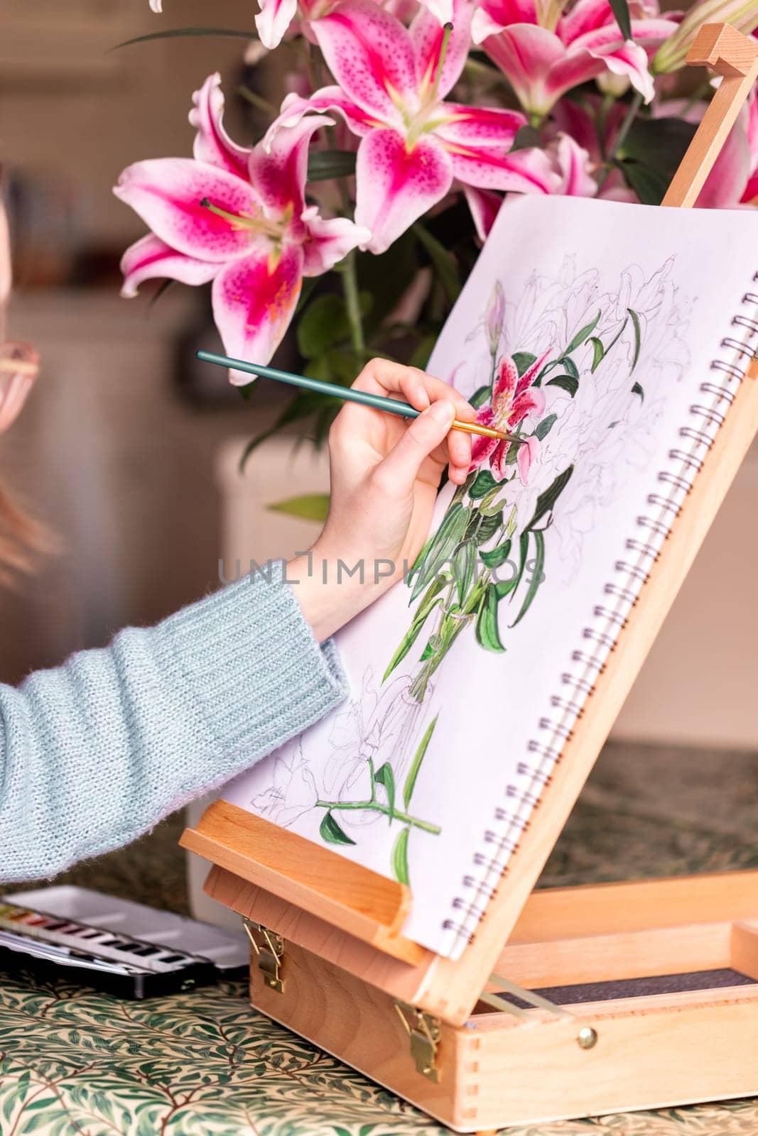 Young left-handed girl painting still life with flowers, purple lilies, with watercolor paints on the easel by Len44ik