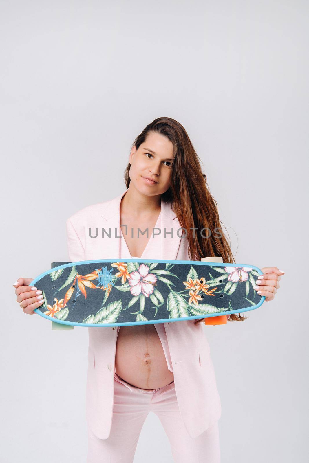 A pregnant girl in a pink suit with a skateboard in her hands stands on a gray background by Lobachad