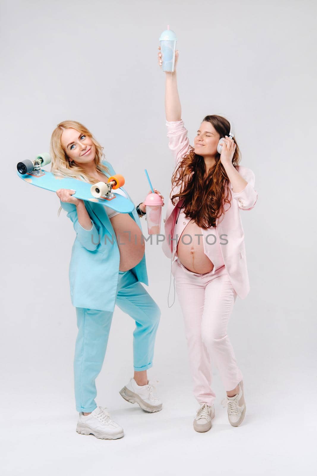Two pregnant girls in turquoise and pink suits with glasses of juice, a skate and headphones stand on a gray background by Lobachad