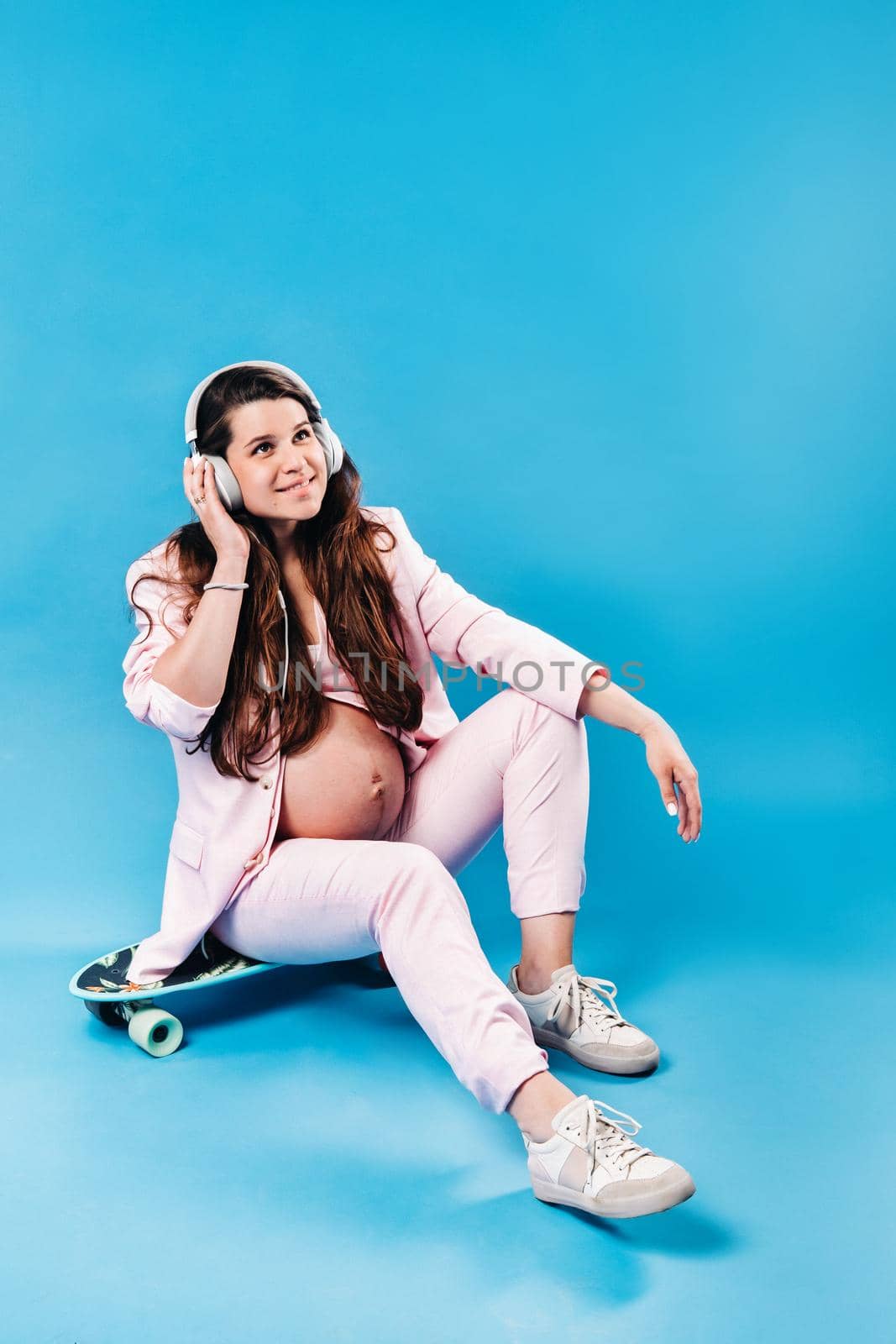 a pregnant woman in a pink suit sits on a skateboard with headphones and listens to music on a blue background by Lobachad
