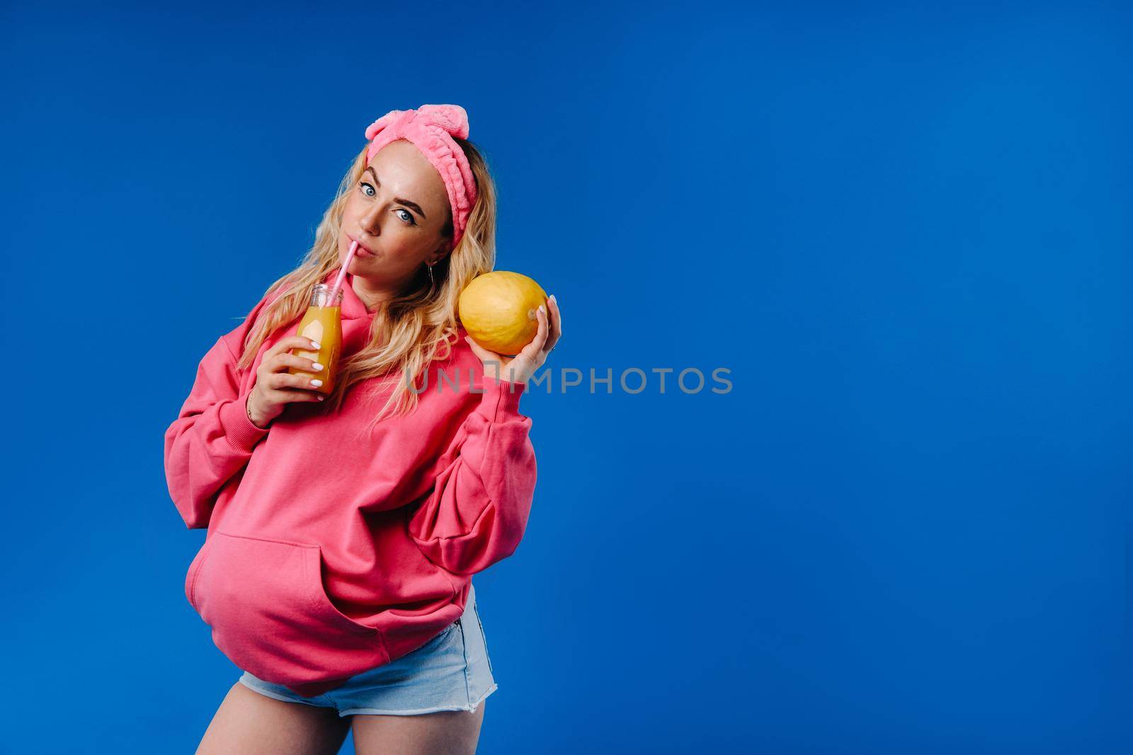 pregnant girl in pink clothes with a bottle of juice and a melon on a blue background.