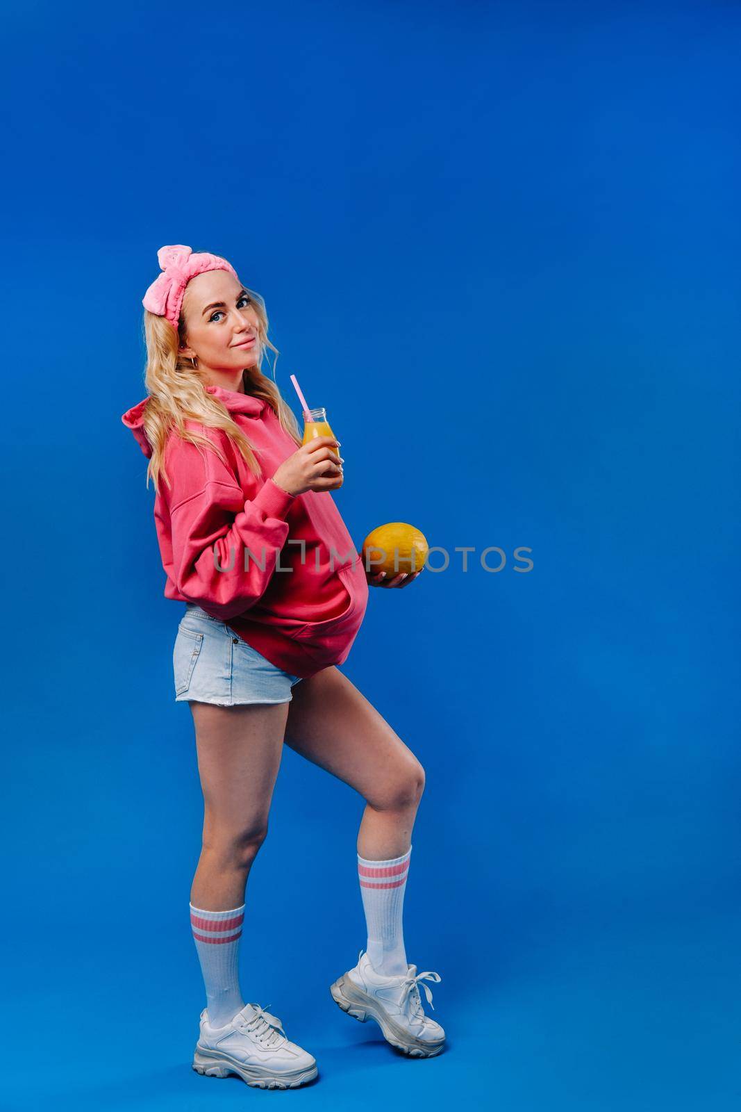 pregnant girl in pink clothes with a bottle of juice and a melon on a blue background.