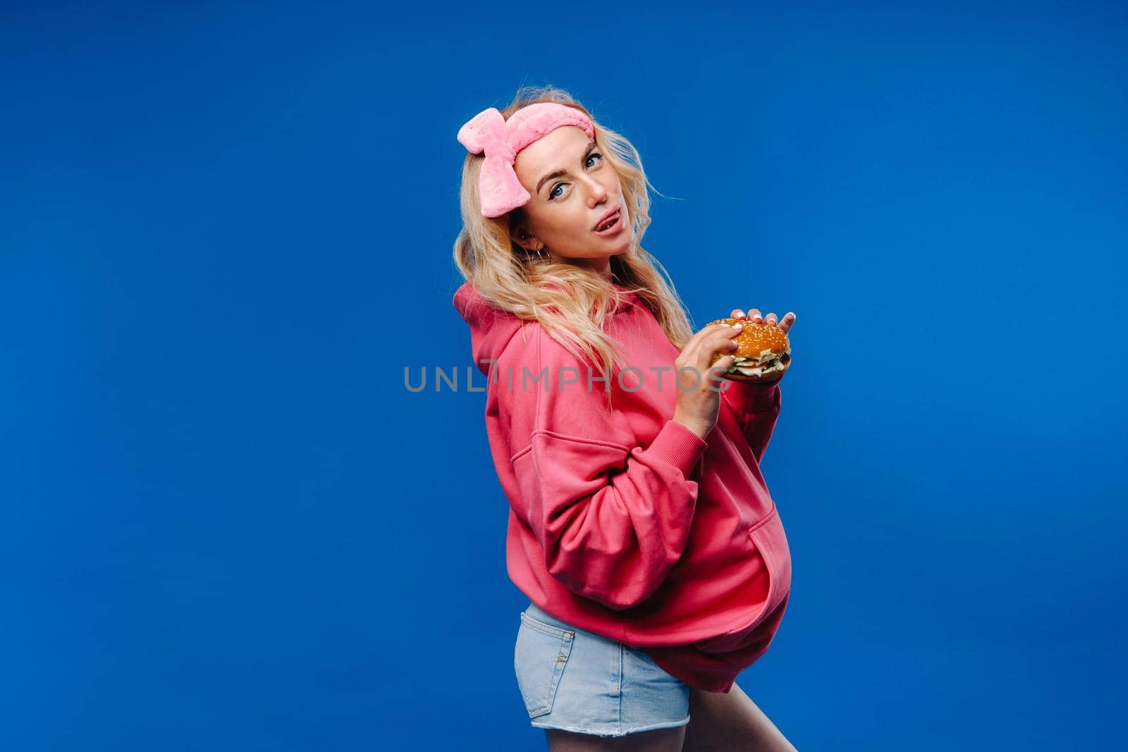 a pregnant girl in pink clothes with a hamburger in her hands on a blue background.