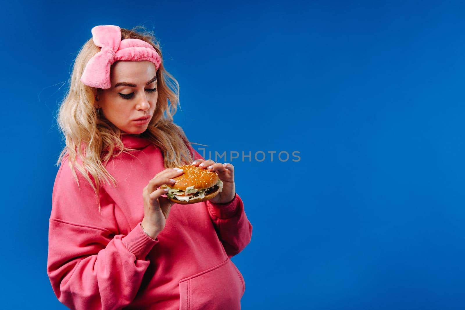 a pregnant girl in pink clothes with a hamburger in her hands on a blue background.