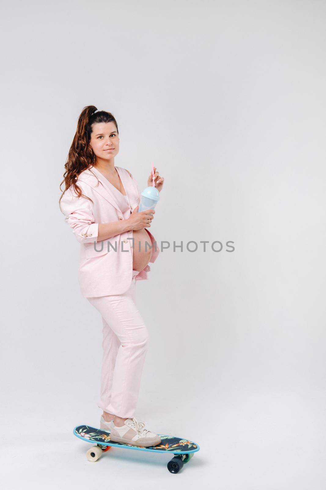 A pregnant girl in a pink suit with a skateboard in her hands stands on a gray background.