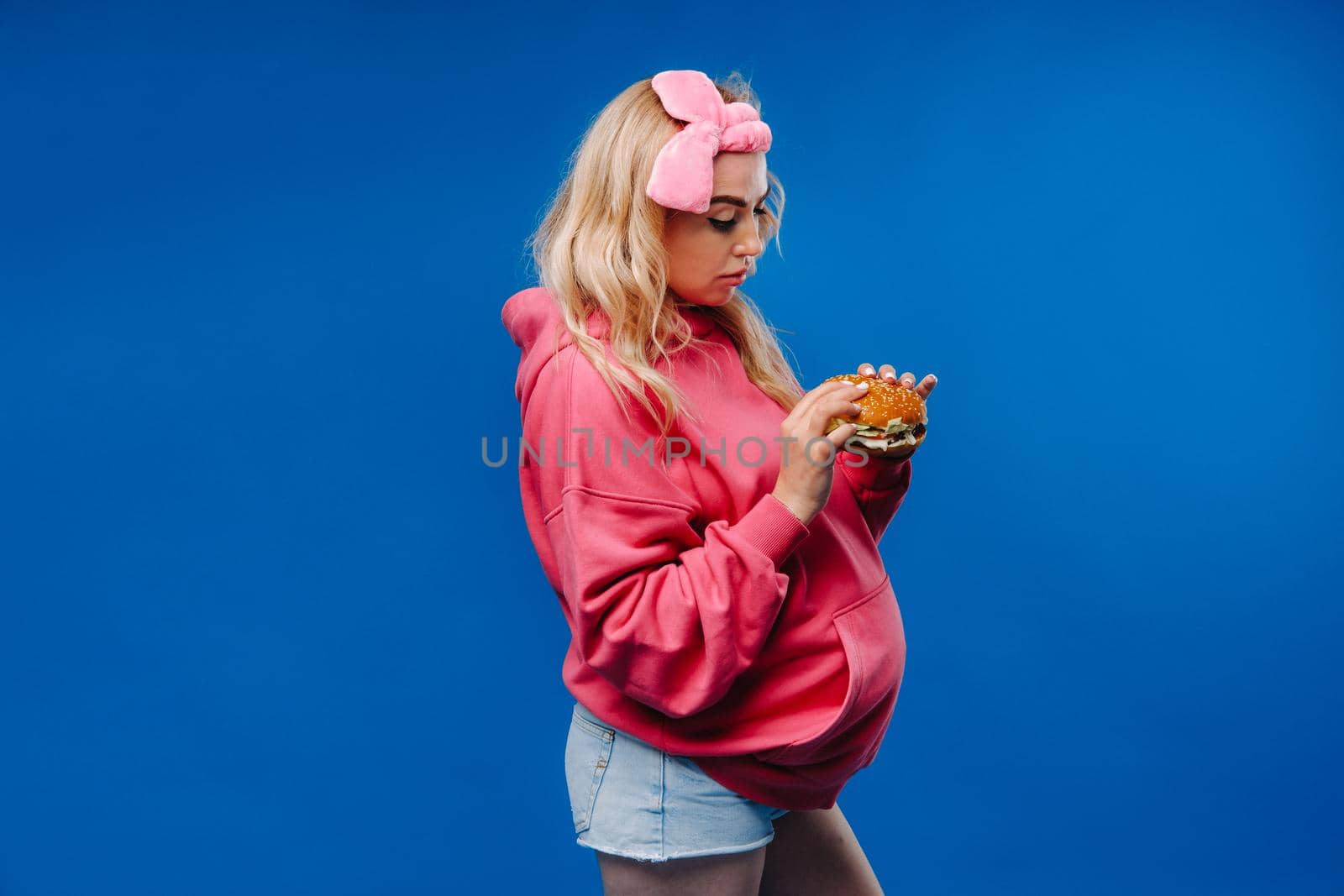 a pregnant girl in pink clothes with a hamburger in her hands on a blue background.