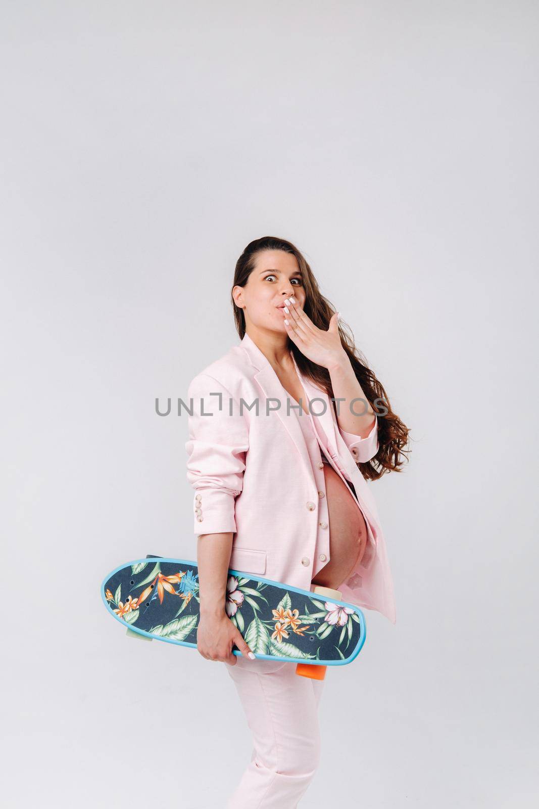 A pregnant girl in a pink suit with a skateboard in her hands stands on a gray background by Lobachad