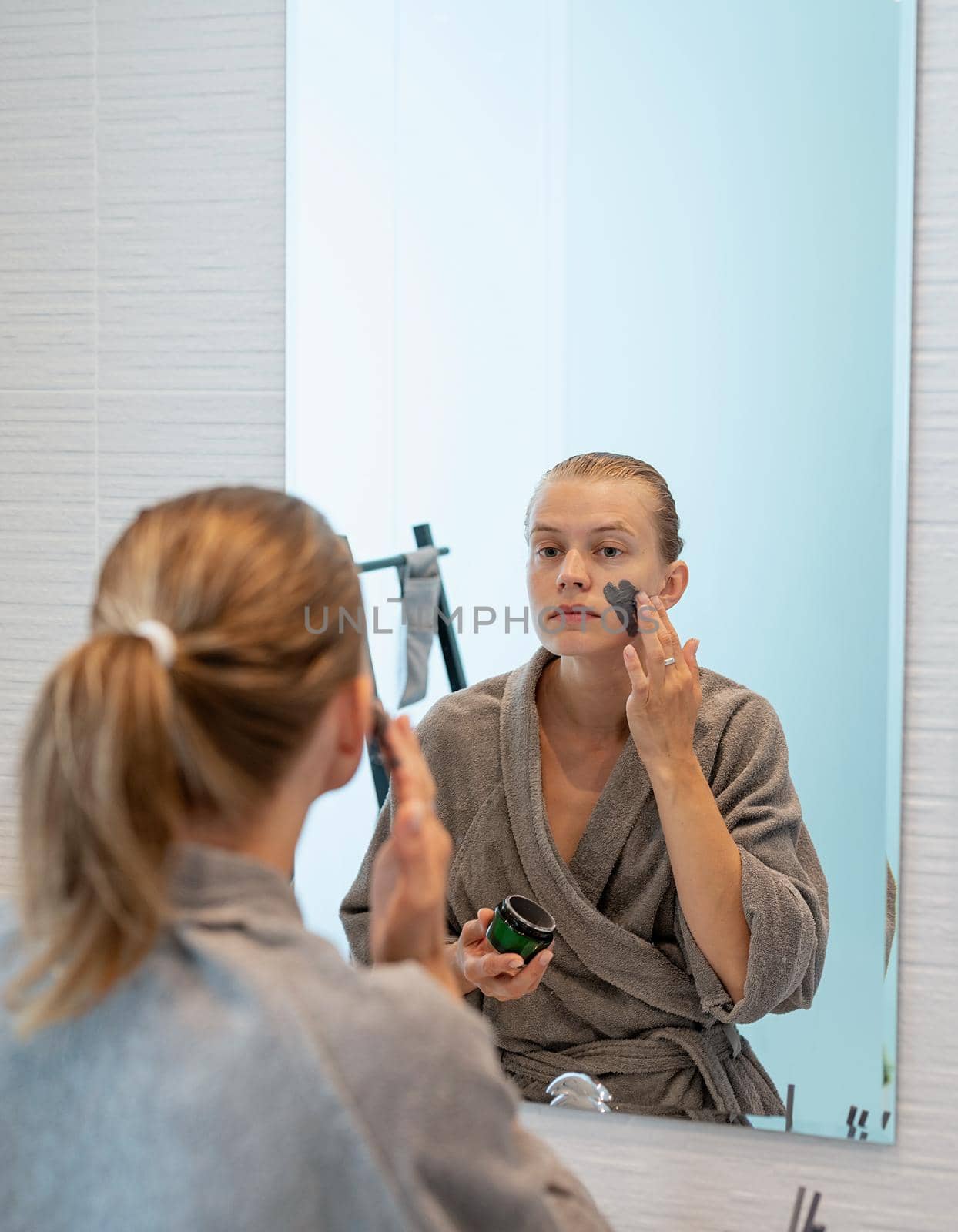 Womens health. Spa and wellness. Woman in gray bathrobes applying face mask in the bathroom looking at the mirror