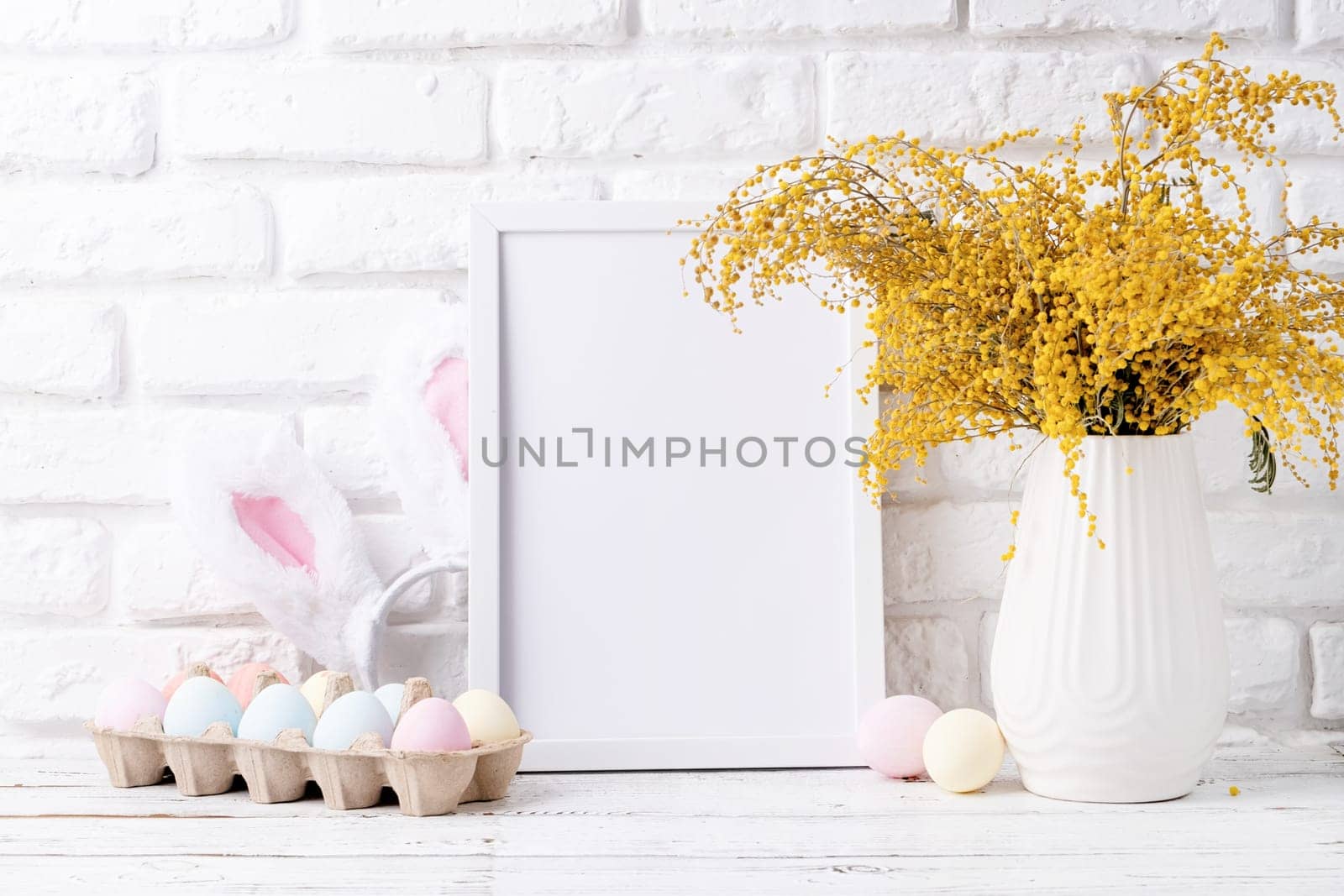 Spring holidays, Happpy Easter. Pastel colored Easter eggs and mimosa flowers with blank white frame for mockup design, front view on white brick wall background