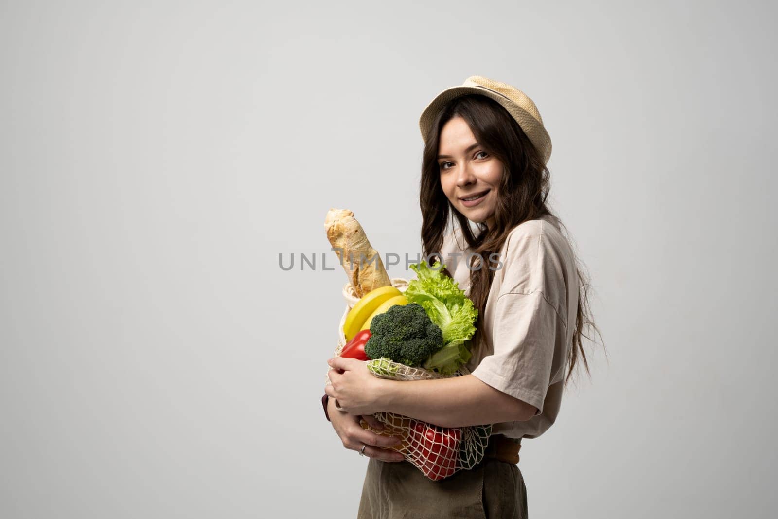 Portrait of happy smiling young woman in beige oversize t-shirt holding reusable string bag with groceries over white background. Sustainability, eco living and people concept. by vovsht