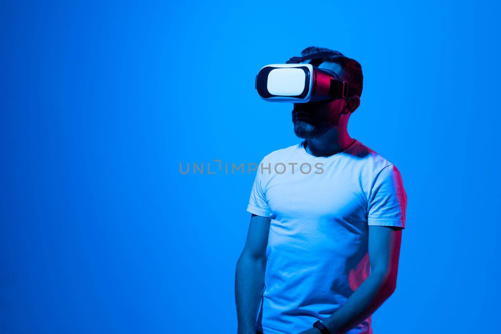 Bearded brunette man in white t-shirt play VR games in virtual reality helmets in neon colourful light. Guy in VR helmets play video games. VR gaming concept. by vovsht